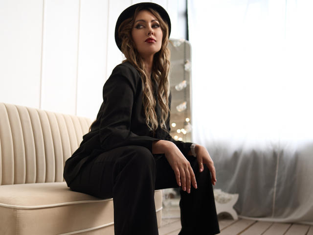 NinaTender, sitting on a beige bench, wearing a black hat, black suit, and black pants, with wavy brown hair, looking to the side, hands on knees.