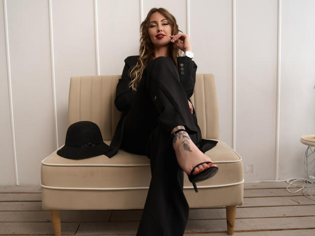 NinaTender with wavy brown hair, black suit, and red heels, sitting on beige couch, right leg up showing tattoo, black hat on left, white wall, wooden floor.
