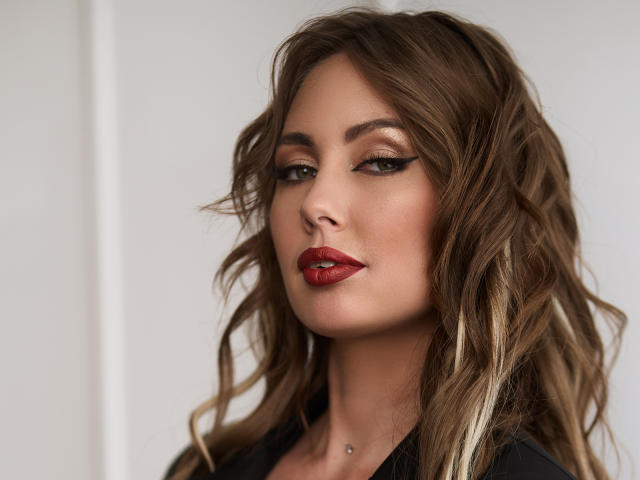 NinaTender with wavy brown hair, red lipstick, and black eyeliner, showing a slight smirk, has a small silver piercing on her lower chin.