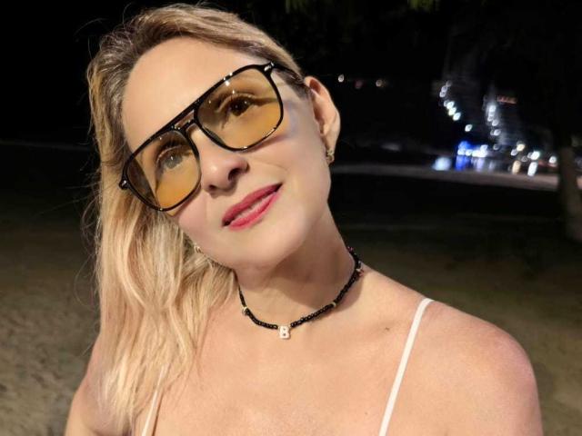 JaneTurner with blonde hair, wearing yellow-tinted glasses, a white spaghetti strap top, and a black bead necklace, smiling at the camera. Night setting, visible city lights.
