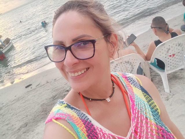 JaneTurner with glasses, smiling, wearing a colorful knit top, necklace, and orange strap, sitting on a beach, holding a phone, showing her tits slightly.