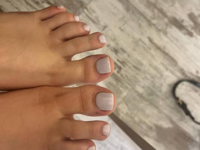 I piedi di Destinis, con le unghie dei piedi dipinte di un colore rosa-grigio chiaro, presentano un aspetto liscio e curato. Le dita dei piedi sono affiancate, con uno sfondo di pavimento in legno..