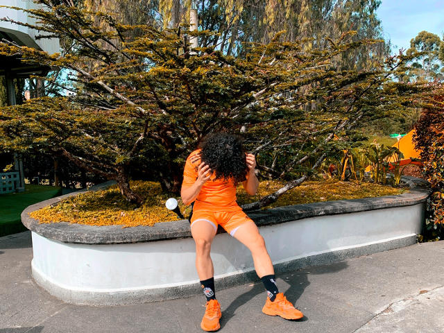 RebelDali69 sits on a curb wearing an orange outfit with matching boots, black socks, and orange shoes, with curly black hair covering their face, in a sunlit park.