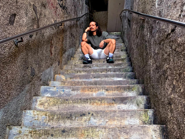 RebelDali69, with medium brown skin, short black hair, and a beard, sits on concrete stairs, legs spread, wearing a gray shirt, white shorts, and black shoes, smiling.