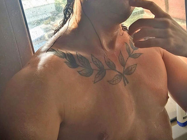 RebelDali69's shirtless upper body, showing a leafy chest tattoo, wet skin, and holding a finger to their lips. Water droplets on their skin. Sunlight from a window.