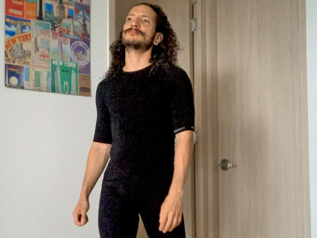 RebelDali69 has long curly hair, a beard, and is wearing a tight black bodysuit, standing with arms relaxed and a slight smirk, in a room with a door.