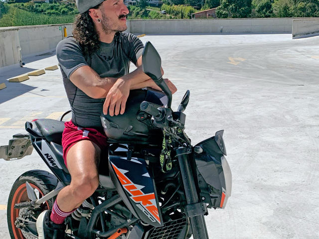 RebelDali69 with long curly hair, gray t-shirt, red shorts, black motorcycle, laughing, wearing a cap, sitting on a black and orange motorcycle, in a sunny parking lot.