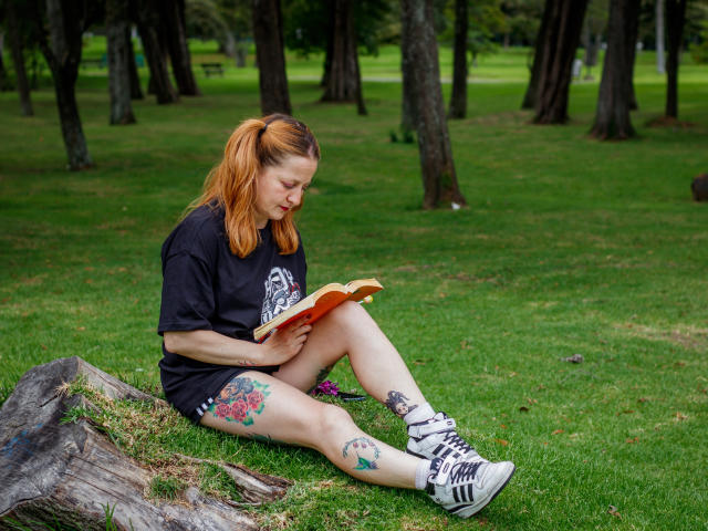 VioletaMora with orange hair, black t-shirt, and shorts, reading a book, has tattoos on legs, white sneakers, sitting on grass, leaning on a log.