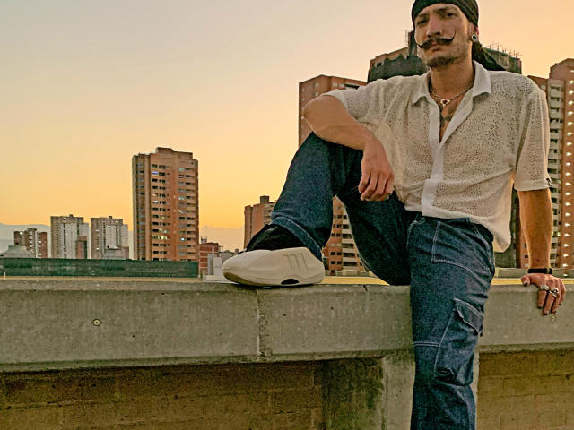 RebelDali69 with a mustache, wearing a white button-up shirt, dark jeans, and white sneakers, sitting on a concrete ledge, one leg raised, city buildings in the background.