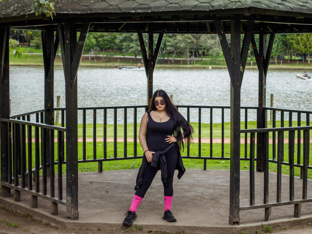 BrithneyLee stands in a wooden pavilion, wearing a black tank top, black pants, pink socks, and black shoes, with long black hair and a confident smile.