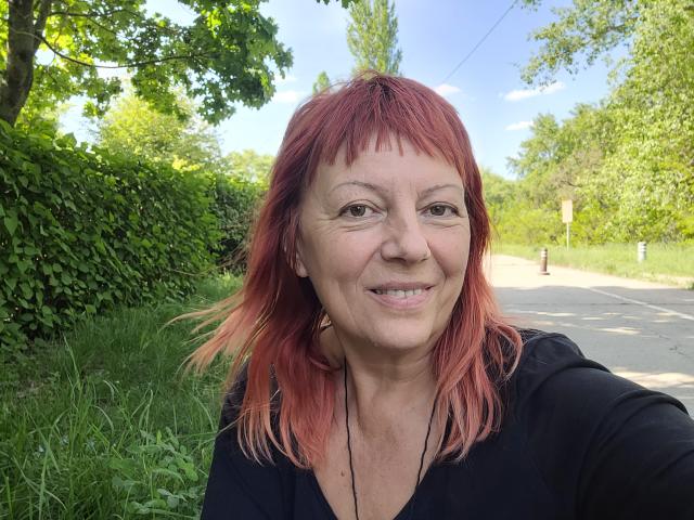 BarbaraBowie has red hair, a black top, and a smiling face with a nose, mouth, and eyes. She's standing on a road with greenery and trees.