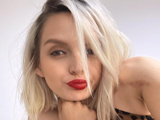 DouceAmelia with blonde hair, red lipstick, and a hand under her chin, looking at the camera, showing her tits in a leopard print top.