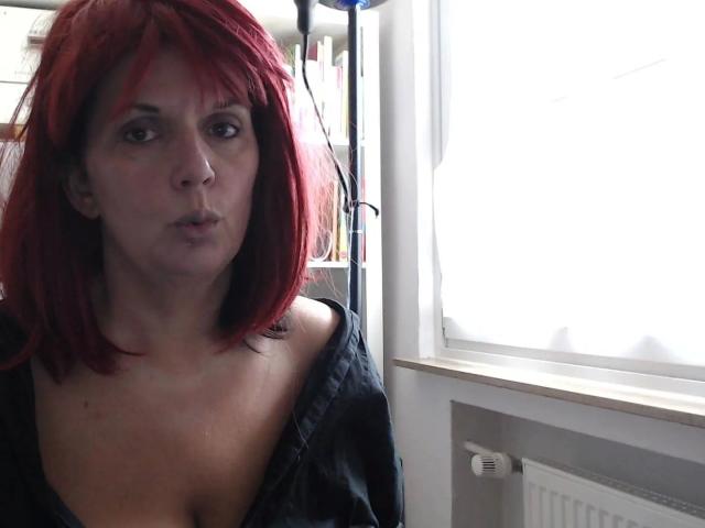 A photo of bigtitsliva-hot with red hair, pouty face, and off-shoulder black top, standing near a bright window, showing a bit of her cleavage.