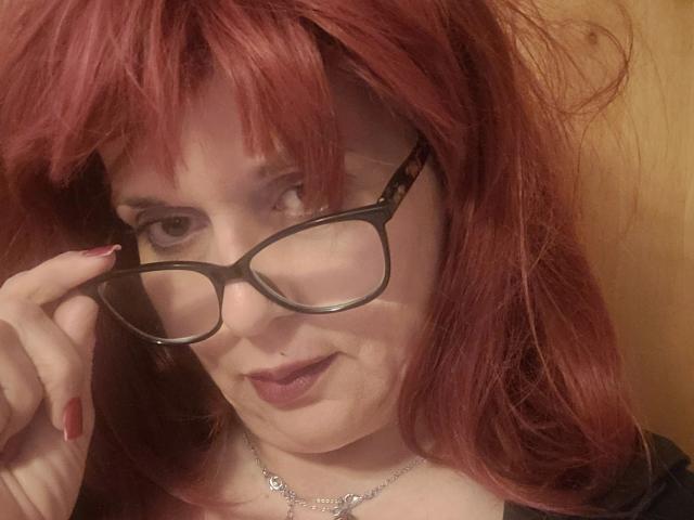 Bigtitsliva-hot with red hair, black glasses, and red nails, wearing a black top and silver necklace, smiling at the camera. Her tits are partially visible.