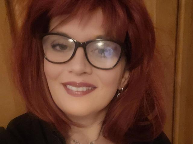 Bigtitsliva-hot with red hair, glasses, and a smile, showing her white teeth and pink lipstick. She's wearing a black top and small silver earrings.