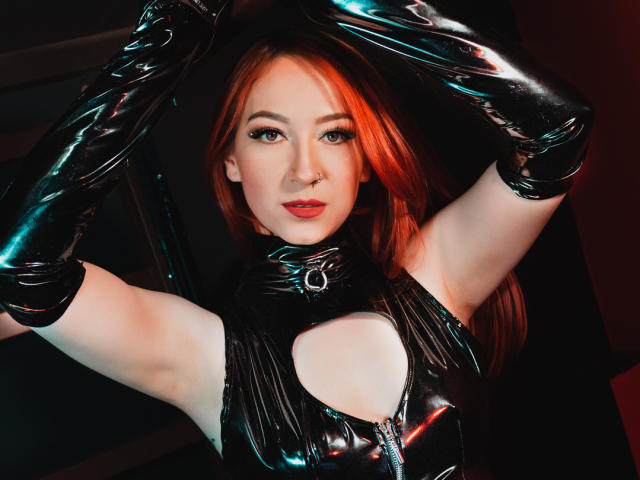 LauraDante with red hair, wearing black latex gloves and cutout top, arms raised, exposing her tits, looking at camera with a nose ring, in a dark, shiny, sexy setup.