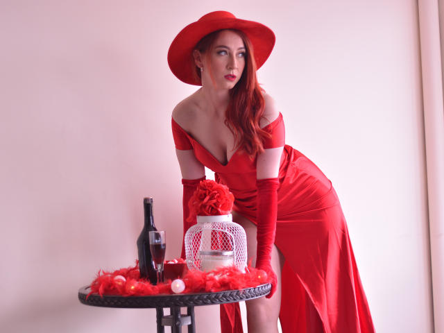 LauraDante com vestido vermelho, chapéu, luvas, apoiada em uma mesa, garrafa, gaiola branca, rosas vermelhas, penas vermelhas, olhos vermelhos, lábios vermelhos, cabelo vermelho, pele pálida, olhando para o lado..