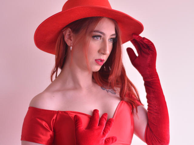 LauraDante with red hat, gloves, and dress, touching her head with red-gloved hand, showing tits with hand on chest, red lipstick, pale skin, red hair, tattoo on right side.