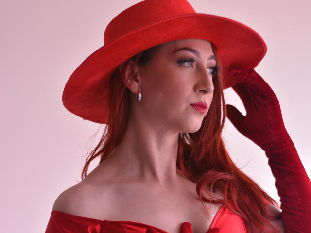 LauraDante with red hat, gloves, and dress, holding hat with right hand, showing off tits, wearing earrings, looking to the side, red hair, pale skin, pink background.
