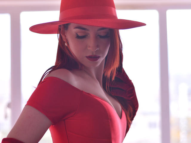 LauraDante com vestido vermelho, blusa com um ombro descoberto, e chapéu vermelho combinando, olhando para baixo, mostrando seus seios, batom vermelho e cabelo longo vermelho, em pé sob a luz forte do sol..
