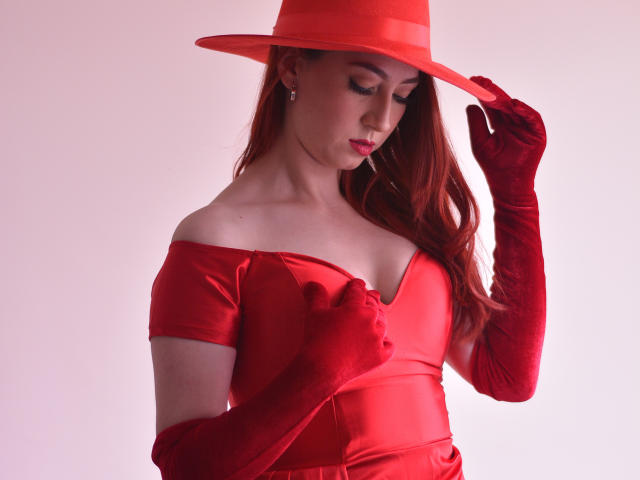 LauraDante com um vestido de veludo vermelho, com um ombro descoberto, e luvas combinando, usando um chapéu vermelho, tocando no chapéu, mostrando os seios, cabelo vermelho longo, pele clara e maquiagem discreta..