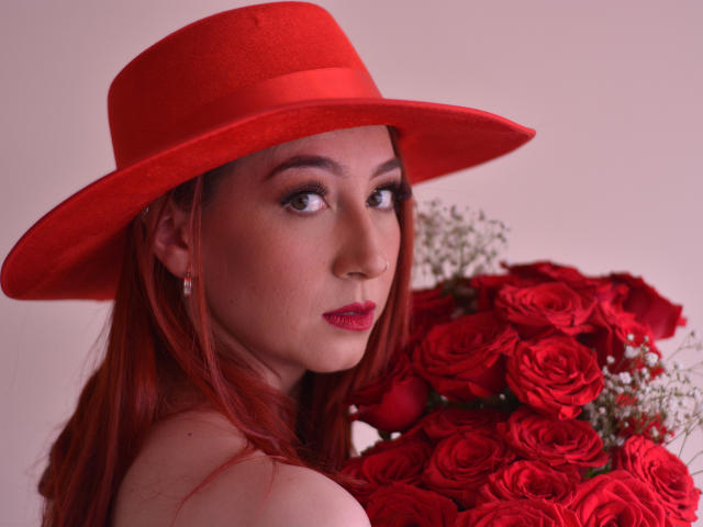 LauraDante com um chapéu vermelho, segurando rosas vermelhas, olhando para trás com olhos sedutores, batom vermelho e brincos, exibindo seus seios e nádegas, com um buquê na mão..