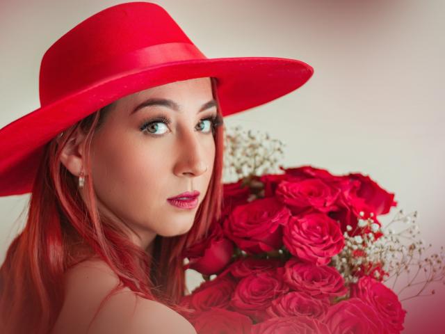 LauraDante with red hat, red lipstick, and green eyes, holding a bouquet of red roses, showing her tits, wearing earrings, and looking at the camera.