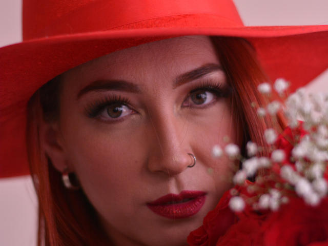 LauraDante com chapéu vermelho, piercing no nariz, batom vermelho e flores vermelhas no rosto, olhos castanhos e cabelo ruivo, olhando diretamente para a câmera. Seus seios estão ligeiramente visíveis..