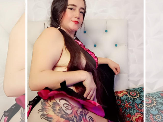 ElizabethSweetyHot with long black hair, pink bra, and black panties, showing a large colorful tattoo on her right thigh, sitting on a white tufted bed. Her red lips are visible.