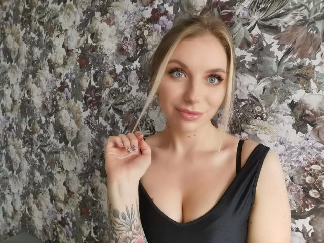SlaviaH-hot with blue eyes, blonde hair, black top, small tattoos, holding hair, showing cleavage, slight smile, pink lips, fair skin, floral patterned wall in background.