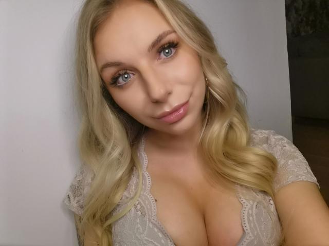 Blonde SlaviaH-hot with blue eyes, wearing a lace top, showing large boobs, has wavy hair, and a slight smile in a selfie.