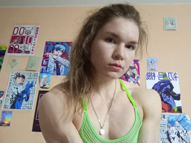 VeraKyk with messy brown hair, green mesh bralette, and pink necklace, showing small tits, and a serious face, surrounded by anime posters on a beige wall.
