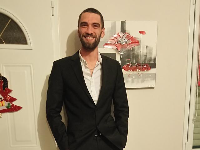 Liam69 is a bearded dude with a black suit, white shirt, hands in pockets, smiling, standing in front of a painting with red flowers.