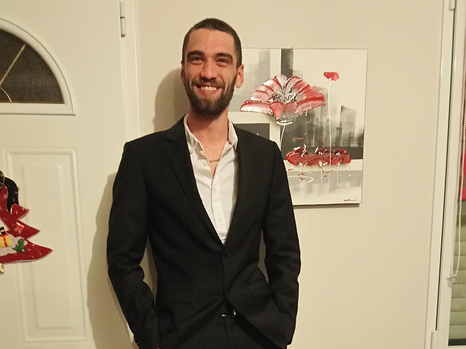 Liam69 is a bearded dude with a black suit, white shirt, hands in pockets, smiling, standing in front of a painting with red flowers.