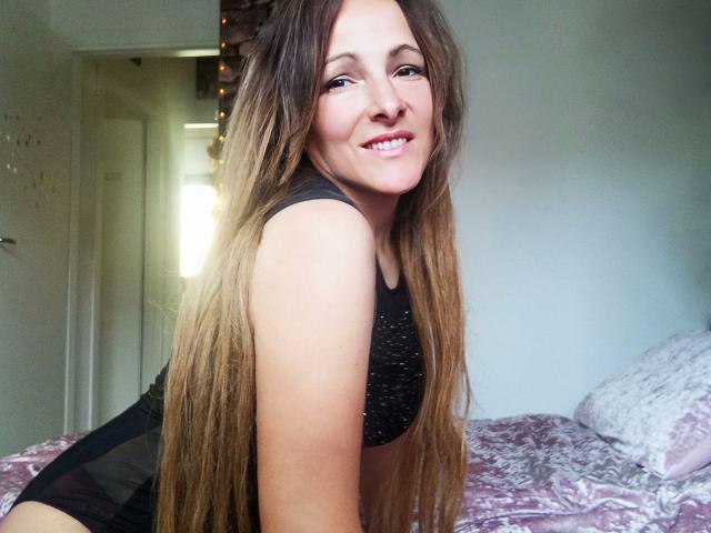 ReifeVivien-hot with long brown hair, smiling, wearing a black sports bra, showing her small ass and cute tits, kneeling on a bed with pink and white patterned sheets.