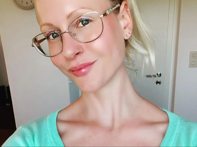 BlondeHexe-hot with glasses, nose piercing, and ear studs, wearing a teal sweater, has pink lips and a slight smile, showing her neck and upper chest.