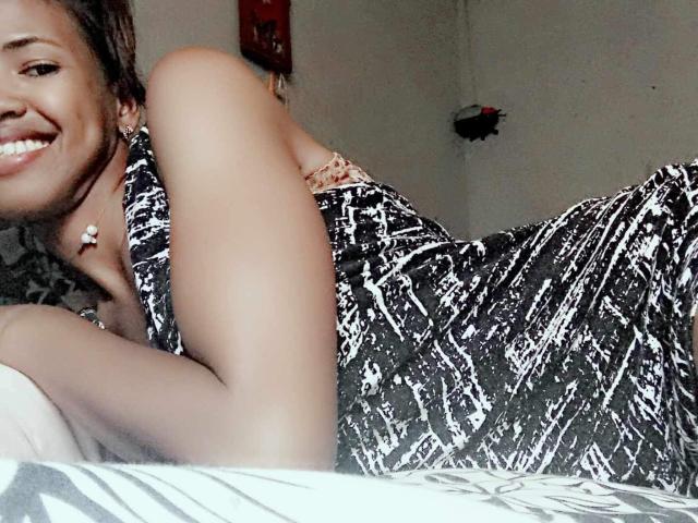 Raynette è sdraiata su un letto, sorridendo, con i glutei in alto e il seno scoperto, indossando un abito a pois bianchi e neri. Ha un orecchino a forma di fiore..