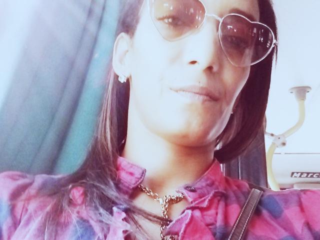 Yay with brown hair, wearing heart-shaped sunglasses, pink plaid shirt, chain necklace, and earrings, smiling slightly, with a bag strap over her shoulder.