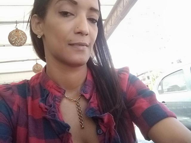 Yay with dark hair, nose ring, and earrings, wearing a red and blue checkered shirt, and a gold necklace, has a small mole under her right eye, and a subtle smirk.