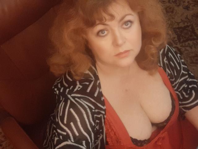 HildaORosse with curly red hair, wearing a zebra-patterned cardigan, red dress, and black lace bra, showing big tits, sitting in a brown chair.