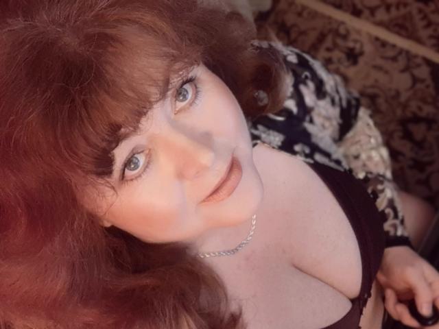 HildaRosse has red curly hair, blue eyes, and pale skin. She's wearing a black bra that shows her big tits. She's looking up with a slight smile.