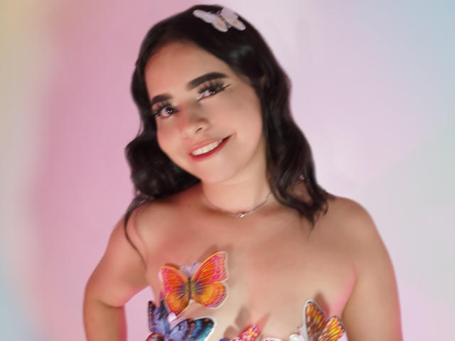 LissyCarter has dark wavy hair with a white butterfly clip, smiling with red lipstick. Her tits are covered in colorful butterfly tattoos, wearing a strapless top.