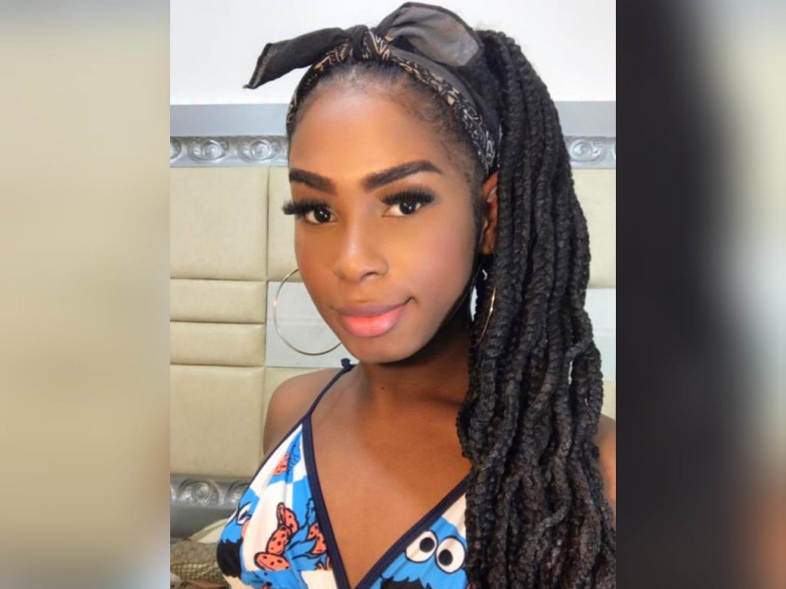 MariposaHornyTs has dark brown skin, long black braids, a black headband, and is wearing a blue, white, and red patterned top. She's smiling with full lips and eyeliner.