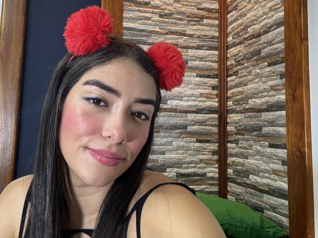 SharonKlart with red fuzzy pom-pom ears, black straight hair, pink lipstick, and a slight smile, wearing a black top, showing her nose ring and subtle makeup.