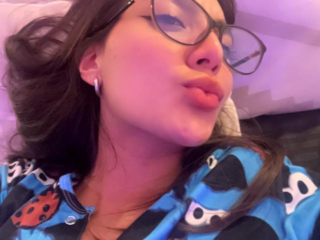 DianaFernandezz pouts with pinkish lips, wearing black glasses, blue pajama top with bug patterns, and white earbuds, her brown hair slightly messy, in a soft purple light.