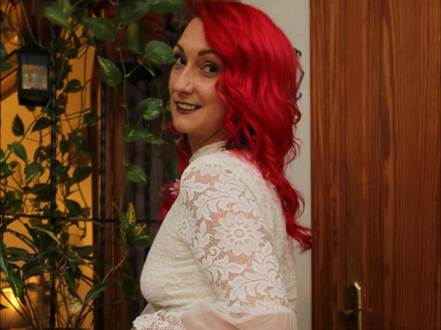 Wild-Vanilla-hot with bright red hair, smiling at the camera, wearing a white lace dress, showing her tits, in a wooden room with a plant and lantern.