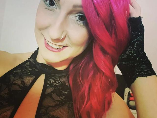 Wild-Vanilla-hot with pink hair, black lace top, and ring, smiling with pierced nose and lip, wearing black lace glove, showing her tits through sheer fabric.