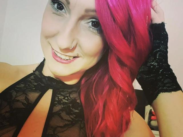 Wild-Vanilla-hot with pink hair, black lace top, and fingerless gloves, smiling at the camera, showing a nose ring and a septum piercing, with visible cleavage.