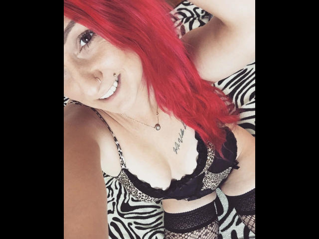 Wild-Vanilla-hot with red hair, smiling, wearing leopard-print lingerie with black lace, has a tattoo under her right breast, and is showing her tits and fishnet stockings.
