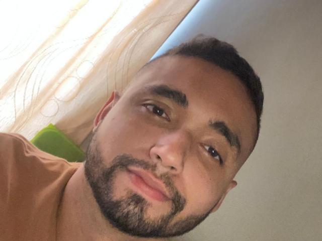 CharlotAndJames has a medium brown skin tone, short curly black hair, a beard, and is wearing a tan shirt. He's looking at the camera with a slight smile.